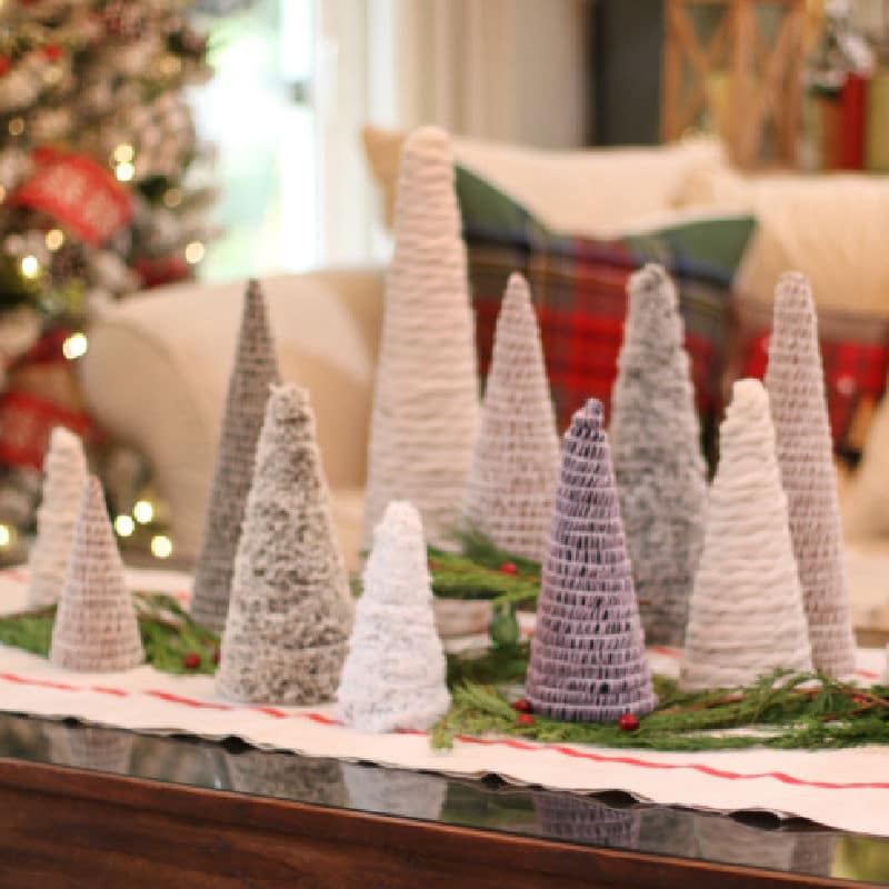 mini Christmas trees of different shapes and textures displayed on bed of evergreens with Christmas tree lights in background