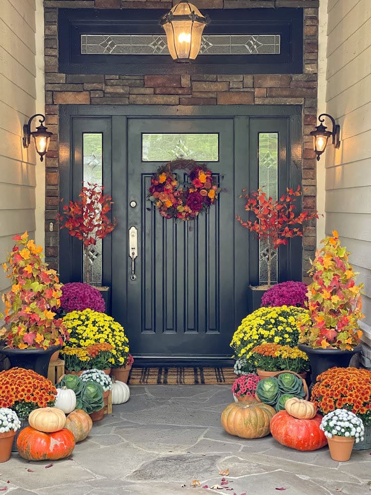 black door exudes sophistication, while a symphony of colorful potted mums in autumnal shades creates a visual delight. A grapevine wreath, carefully woven with seasonal accents, graces the door with its natural charm. Pumpkins in various sizes welcome visitors to bask in the beauty of the season.