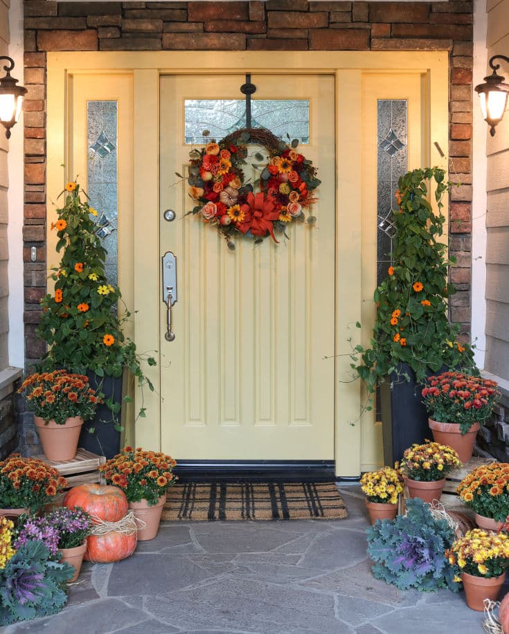 A creamy yellow door and plaid door mat are flanked by two tall trees. This front porch is also surrounded by potted mum, purple kale and colorful pumpkins