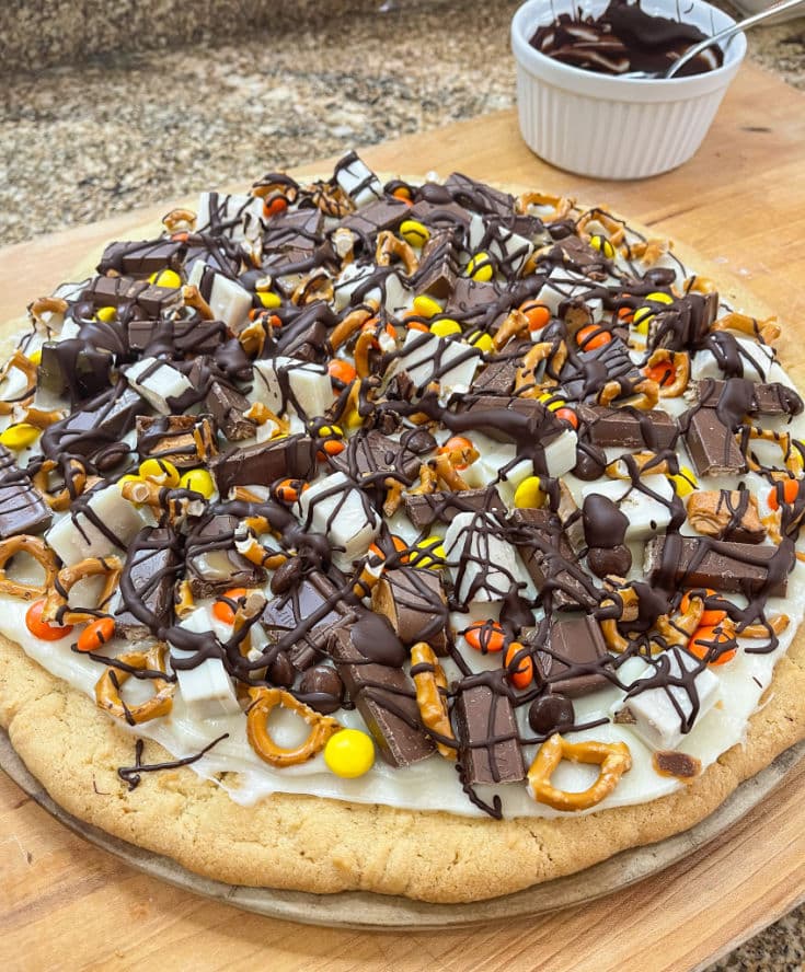 A close-up of a Dessert Pizza, showcasing a sugar cookie crust covered with a colorful array of leftover Halloween candy pieces and drizzled with milk chocolate.